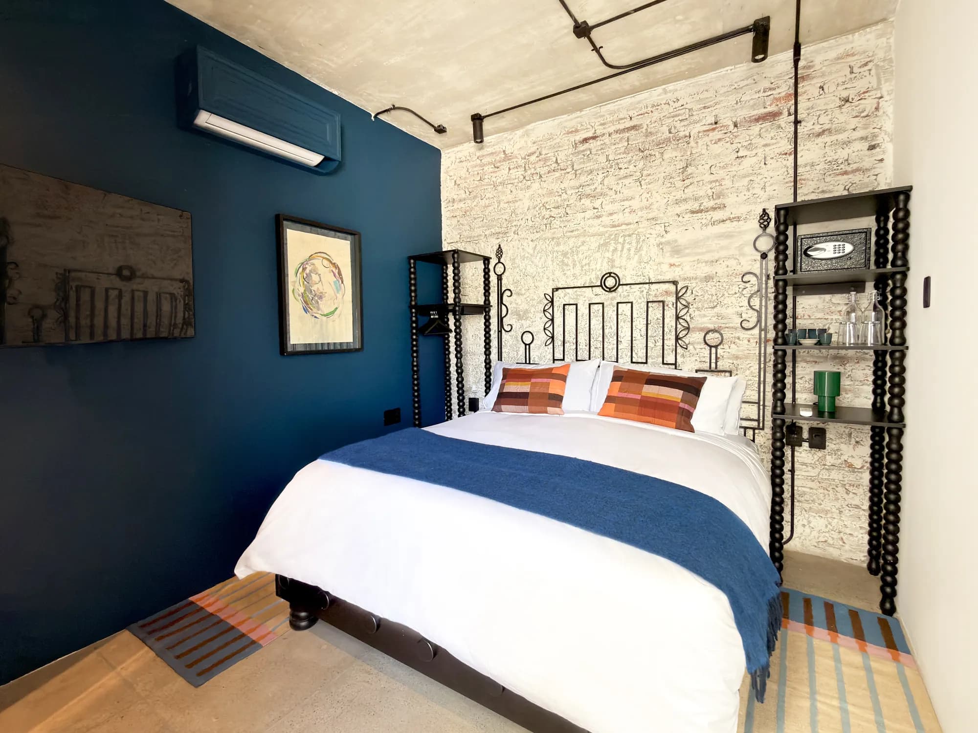 The King bed against the whitewashed brick wall and the petrol-blue wall, with the black shelf of turned wood