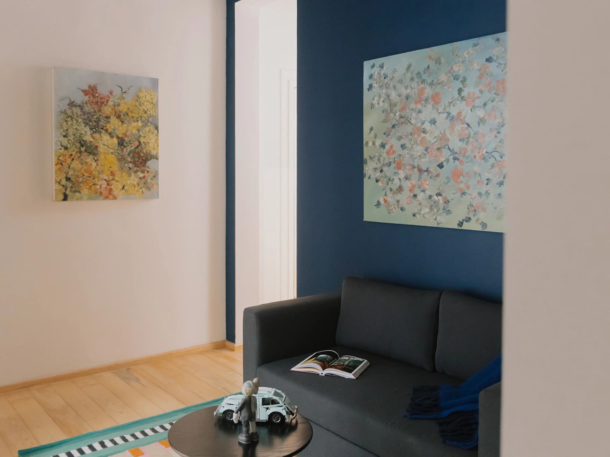 The living area: a grey sofa against the blue wall under floral oil paintings, with a round table of turned wood and a toy car