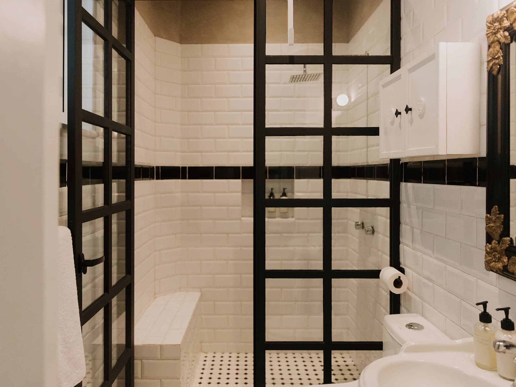 The bathroom with a shower behind glass with black ironwork and white tiled walls