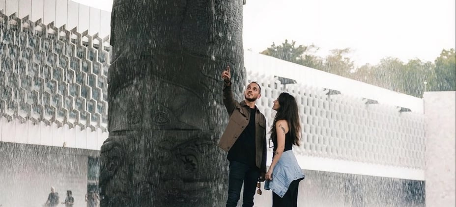 A couple under the "El Paraguas" rain, reaching to touch the water falling from the carved column at the National Museum of Anthropology