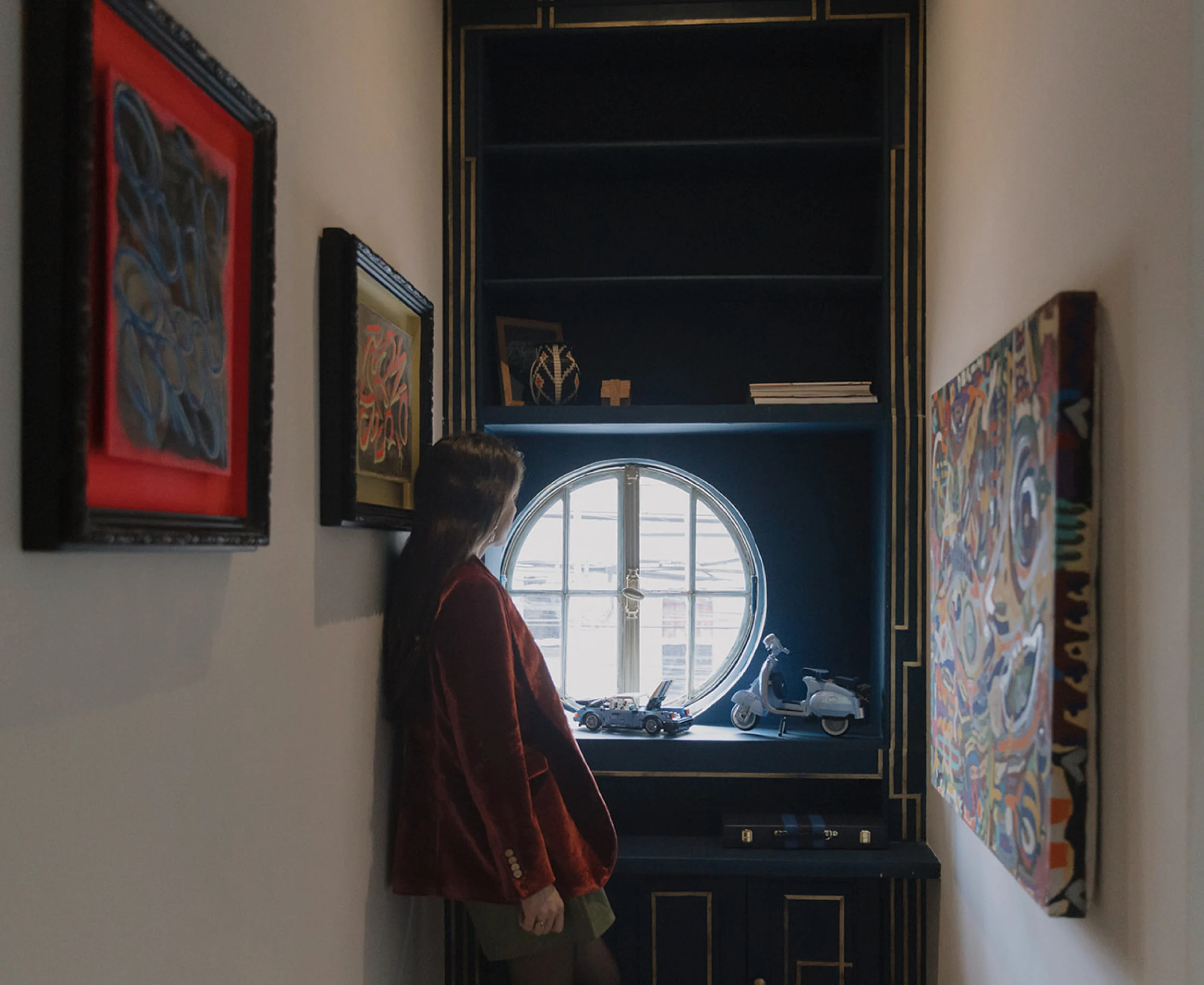 A guest in a red blazer looks at the paintings in a hallway, beside a round window