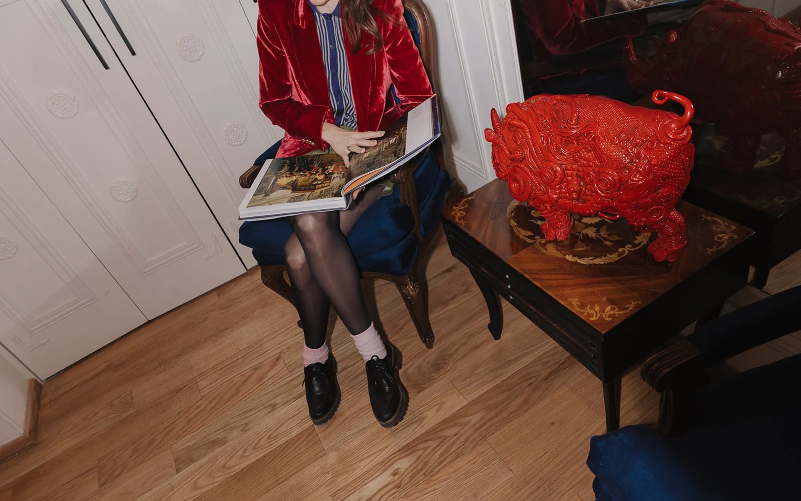 A guest in a red blazer leafs through an art book, seated on a blue velvet chair beside a small table with a red lacquered sculpture