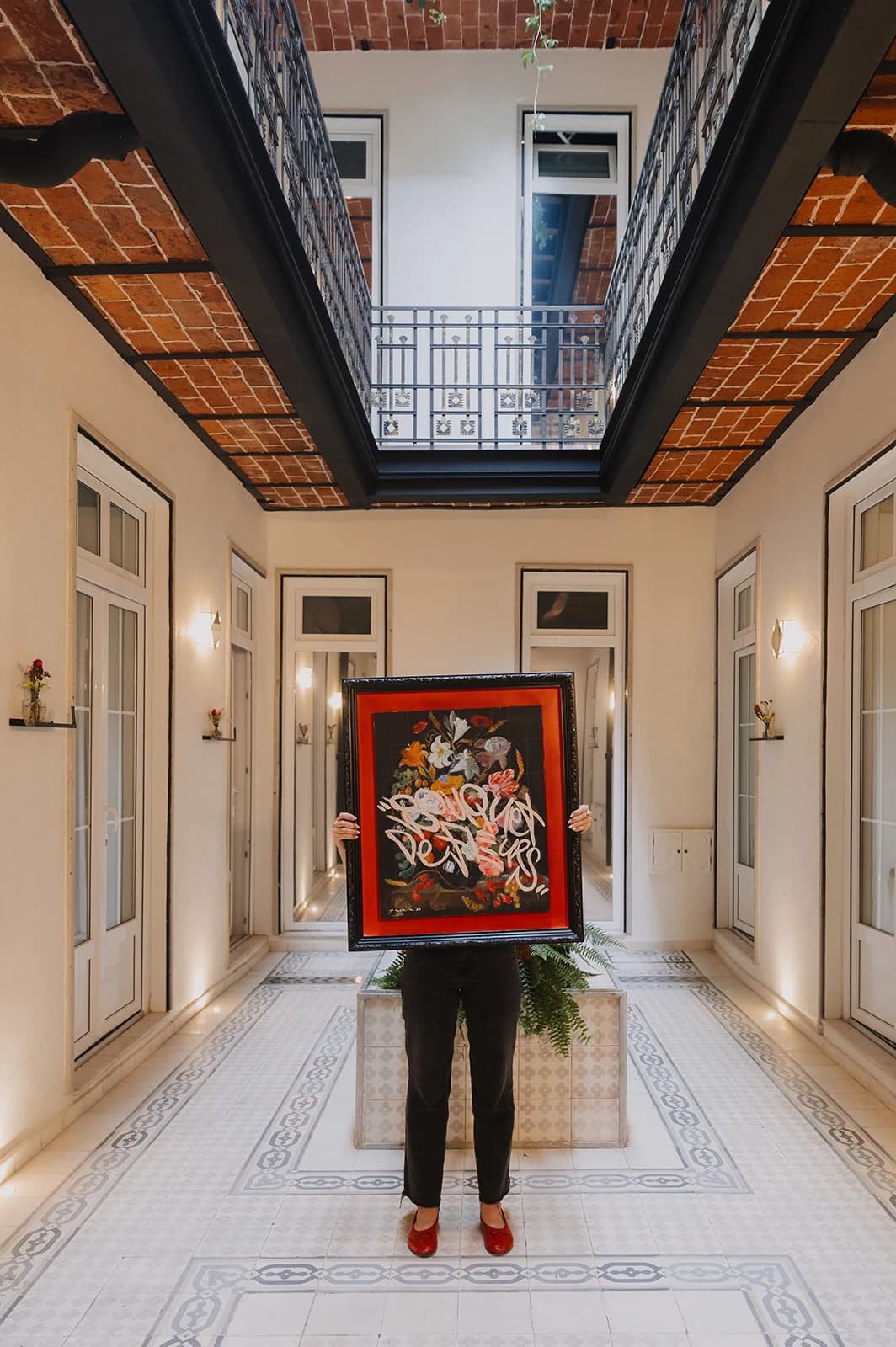 A person in black trousers holds up a red-framed painting of flowers in the house's corridor, with its mosaic floor and brick ceiling