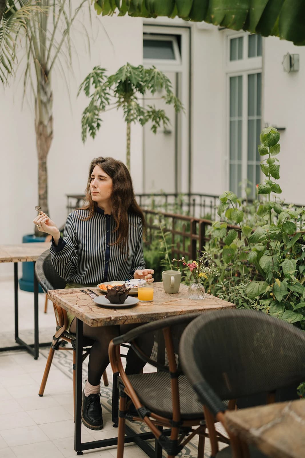 A guest in a striped blouse has breakfast at a wooden table on the garden terrace, among plants and large white windows