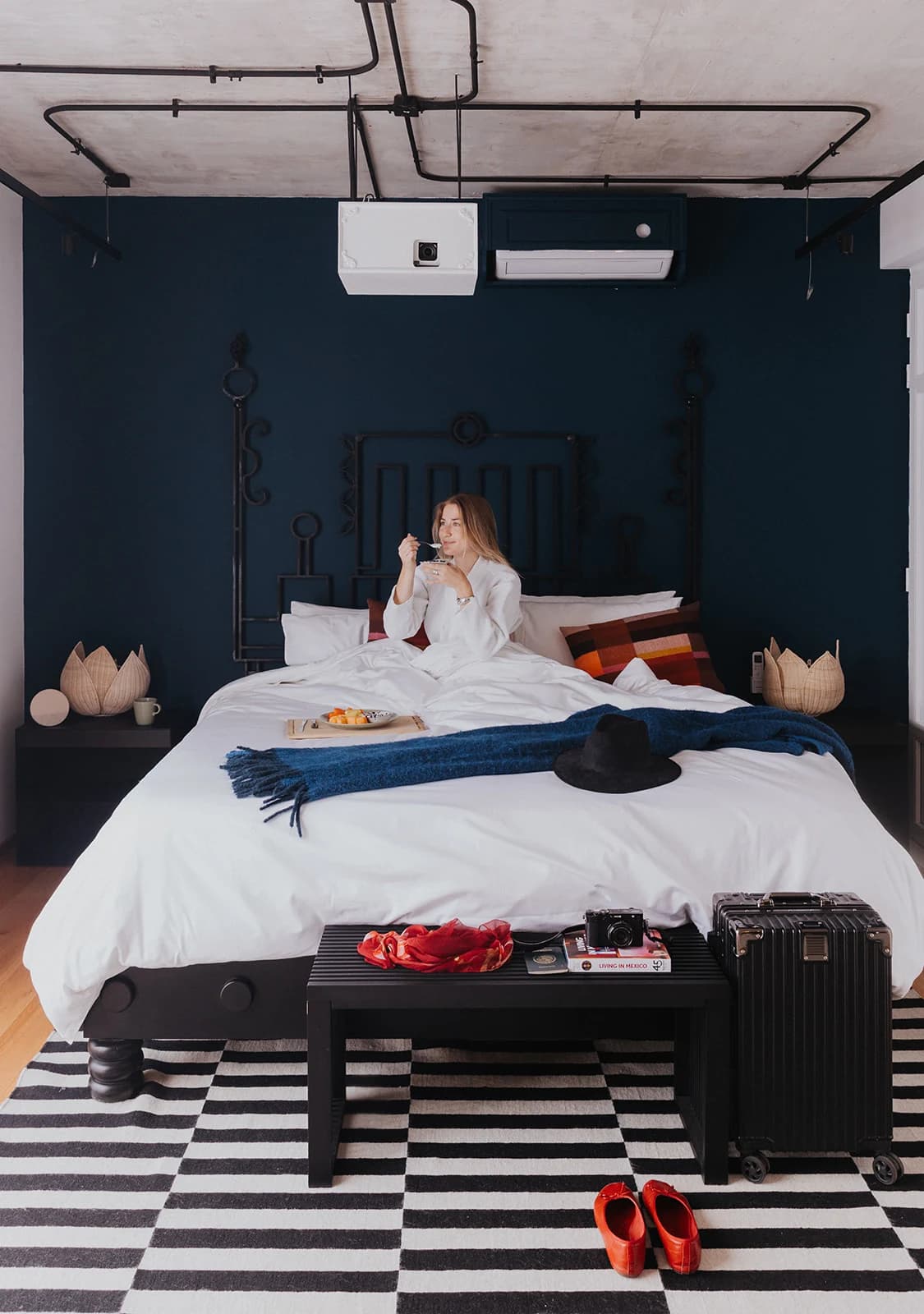 A guest in a robe has breakfast on a king bed with a wrought-iron headboard, in front of a navy wall, with a suitcase and red shoes on the black-and-white checkered floor