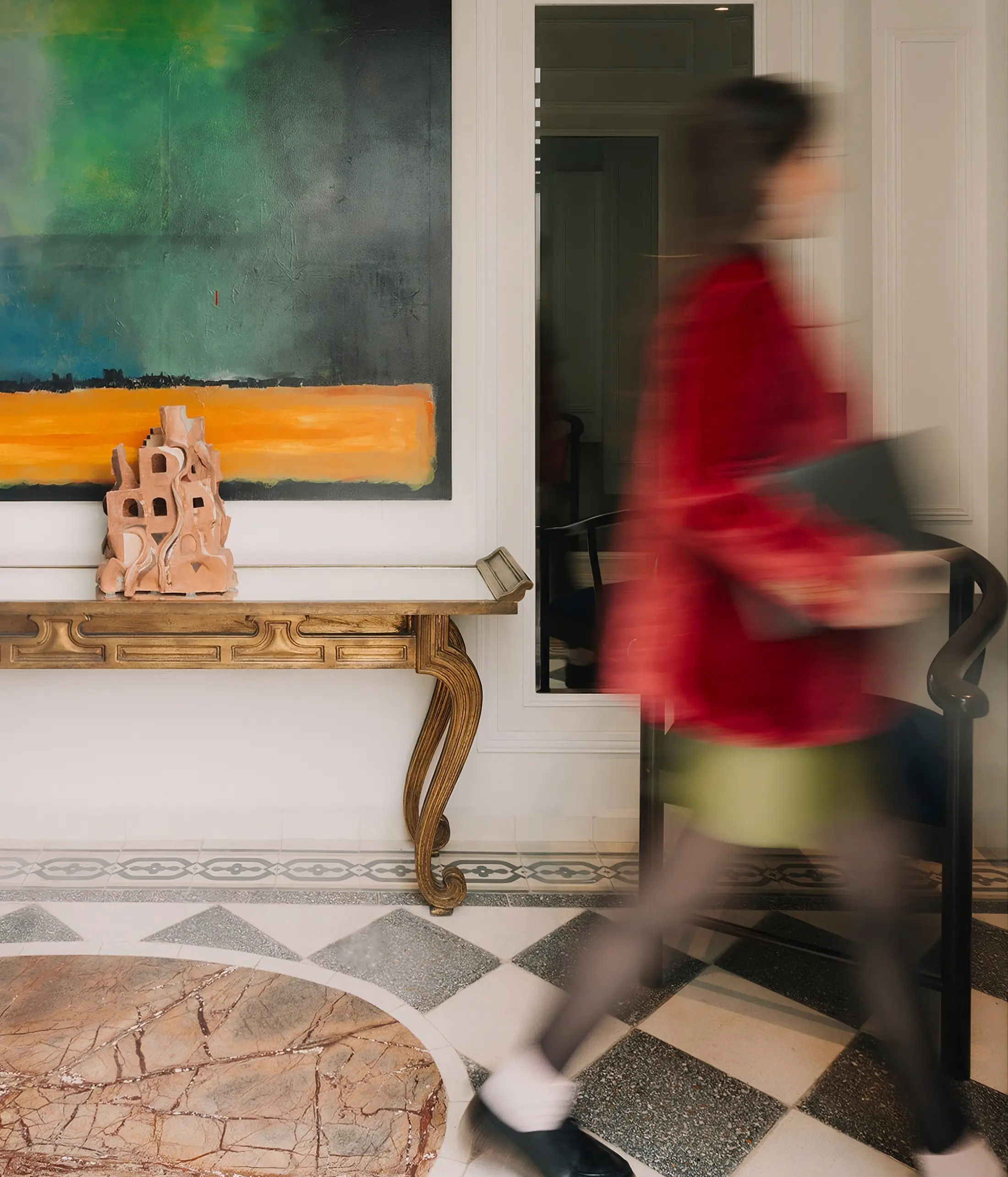 A guest in a red blazer crosses the lobby in motion, beside a gold console and a green painting