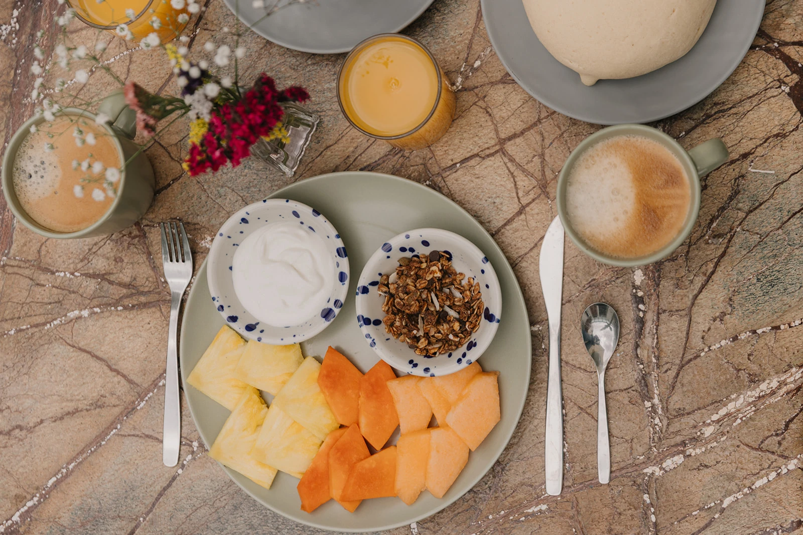 Breakfast seen from above: fruit, yoghurt and granola, a latte, orange juice and bread on veined marble