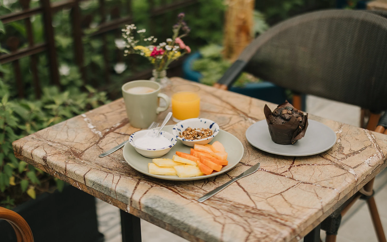 The breakfast table among the terrace plants, with coffee, juice, fruit, granola and a chocolate muffin