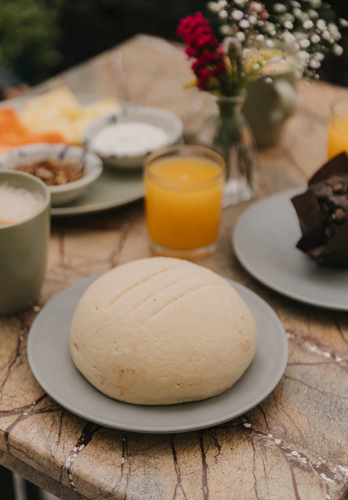 A sweet bread on a green plate, with orange juice and colourful flowers on the marble table