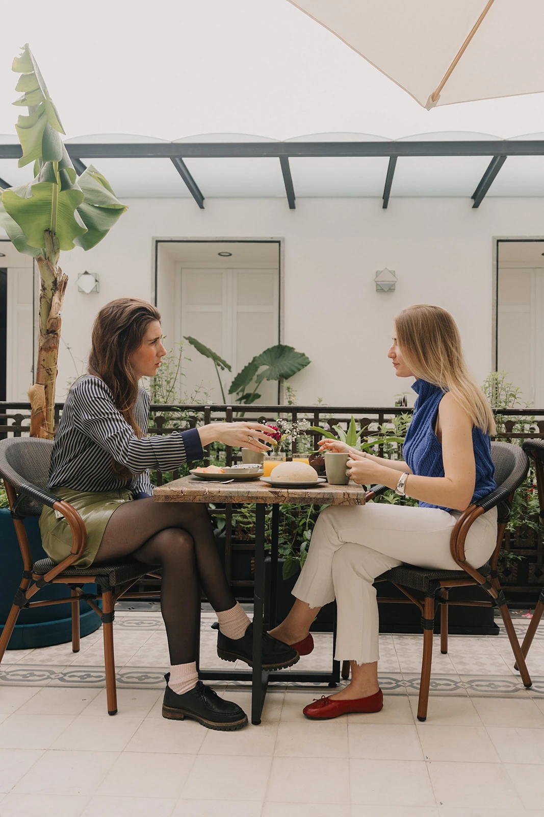 Two guests chatting and having breakfast face to face on the garden terrace, under a parasol