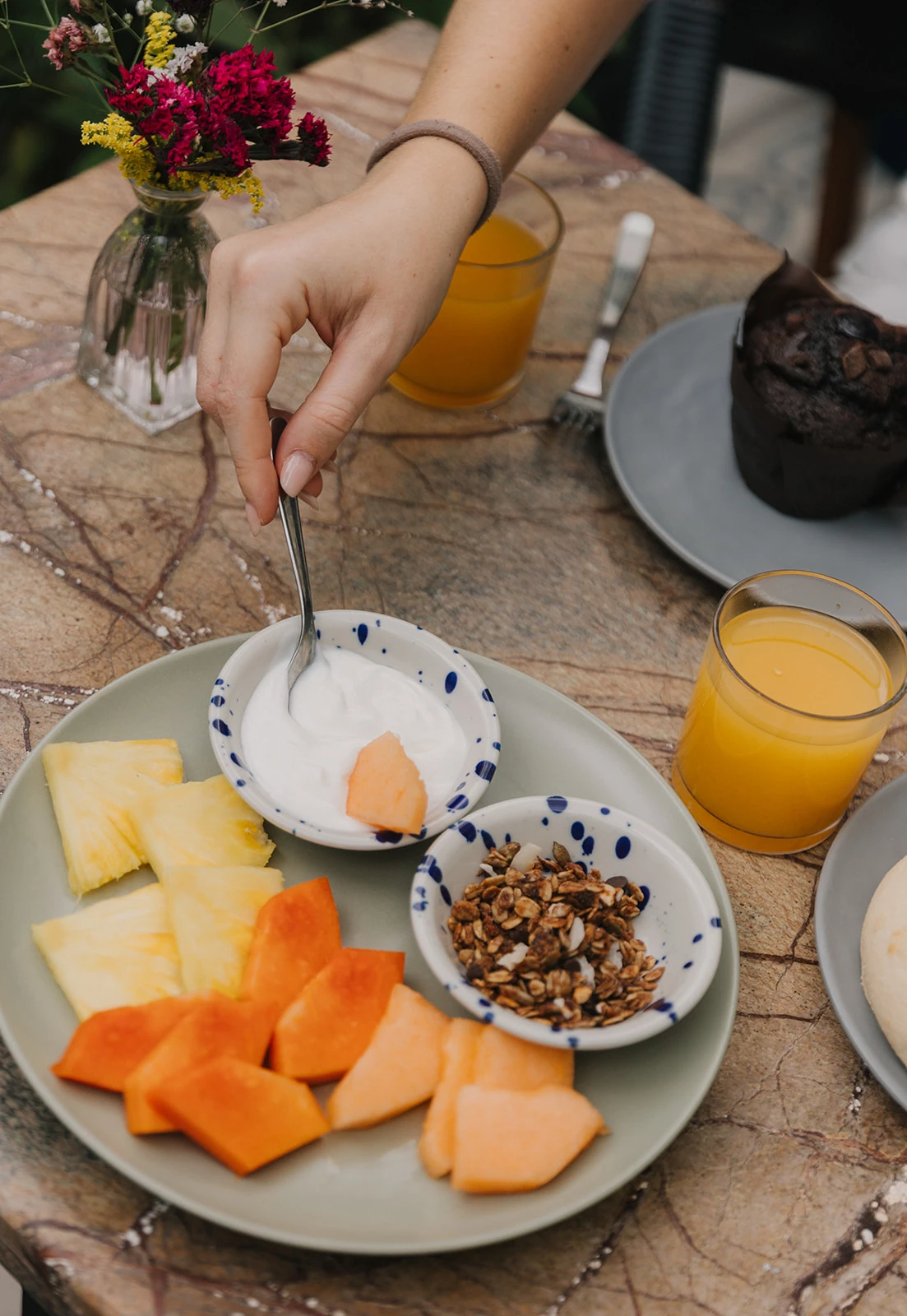 A hand pouring yoghurt over fruit, beside the granola, the juice and a vase of flowers
