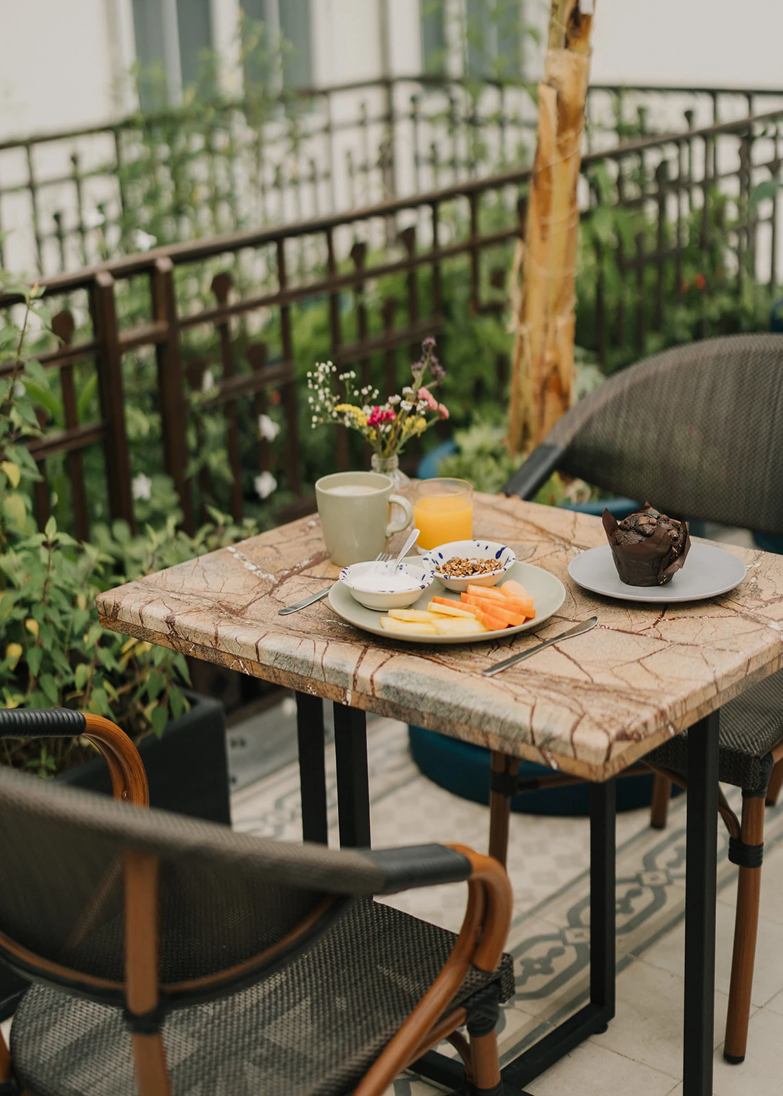 The breakfast table among the terrace plants, with coffee, juice, fruit, granola and a chocolate muffin