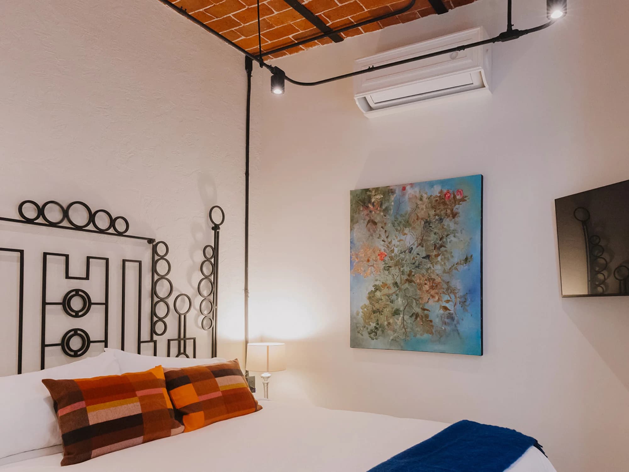 The King bed under the brick-beamed ceiling, with colourful cushions, a floral oil painting and the air conditioning