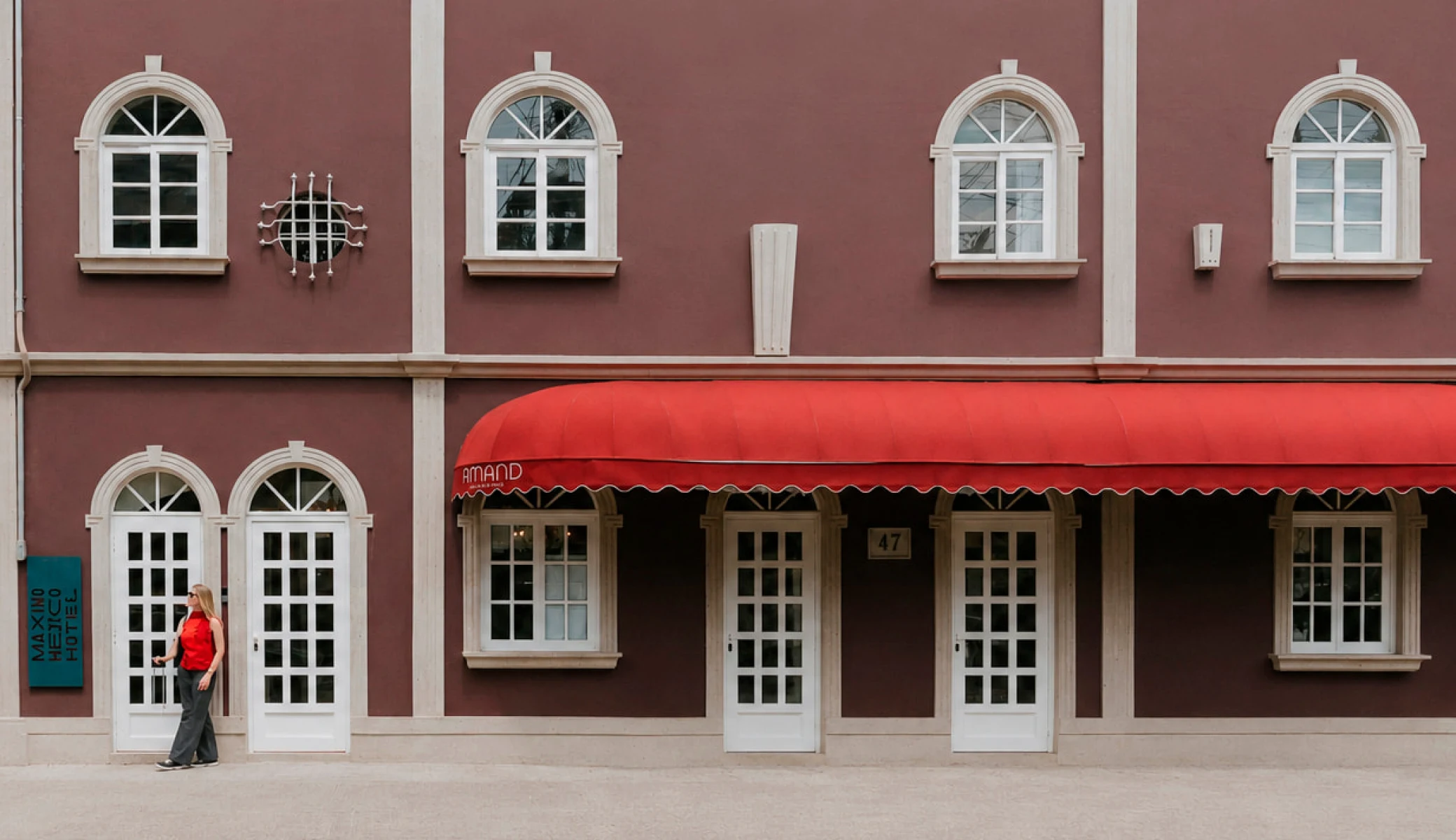 The facade of Máximo México Hotel on Calle Manzanillo: a wine-coloured wall with white arched windows, the house's turquoise vertical sign, Amand's red awning, and a guest arriving with a suitcase