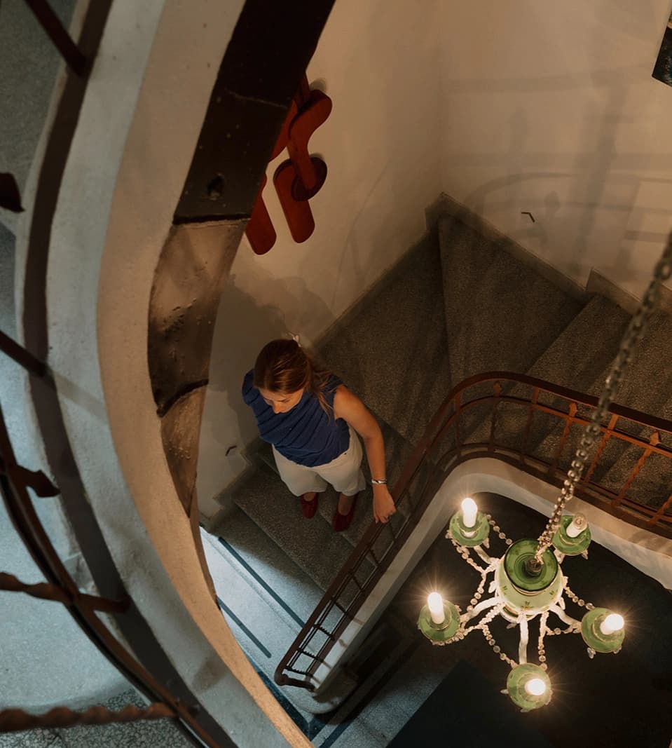 The curved staircase seen from above, with a guest coming down and the green glass chandelier hanging in the well between the wrought-iron banisters