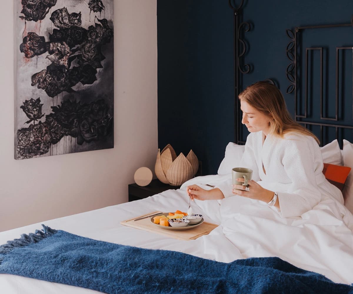 Someone in a white robe having breakfast in bed from a tray, beside a blue wall and a painting of dark flowers